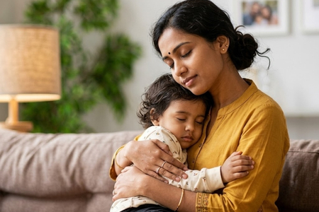 An Indian mother gently cuddles her sleeping toddler on a sofa at home, offering comfort and a sense of security.