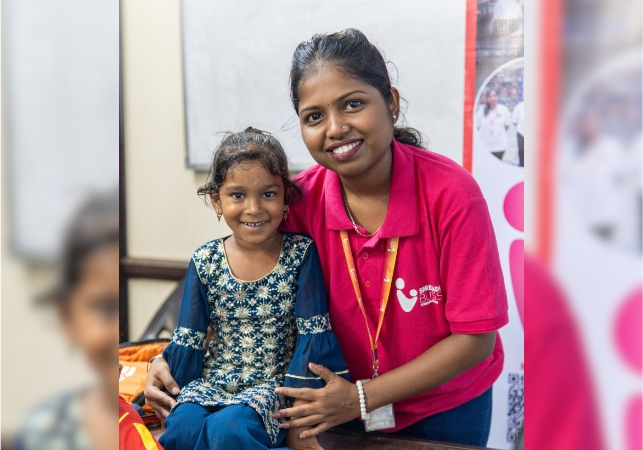A Spreading Bliss Foundation volunteer in a pink polo shirt poses with a young girl at an indoor centre, smiling warmly at the camera.
